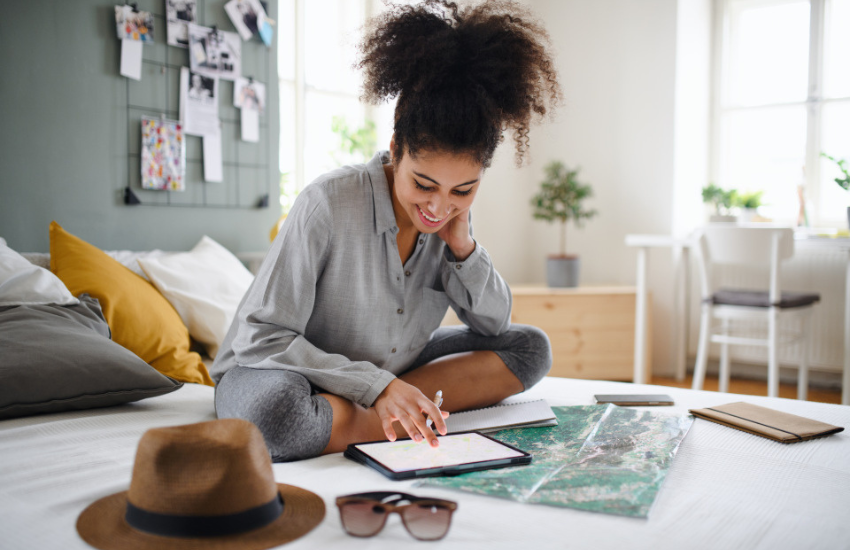 Personne préparant un voyage, consultant une tablette avec une carte et des documents de voyage disposés sur un lit, dans une chambre lumineuse.